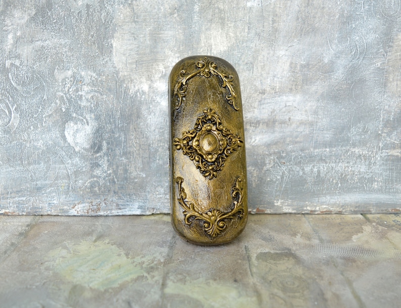 May include: A gold-colored decorative box with ornate floral designs and a raised circular center piece. The box is made of a textured material and has a slightly aged appearance.