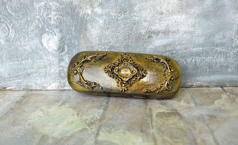 May include: A gold-colored, ornate eyeglass case with a diamond-shaped design in the center. The case is made of a hard material and has a smooth, polished finish.