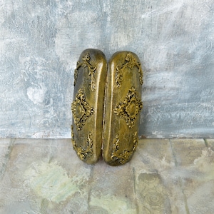 May include: A pair of ornate gold-colored boxes with intricate floral designs and a raised center piece. The boxes are closed and sitting on a textured surface.