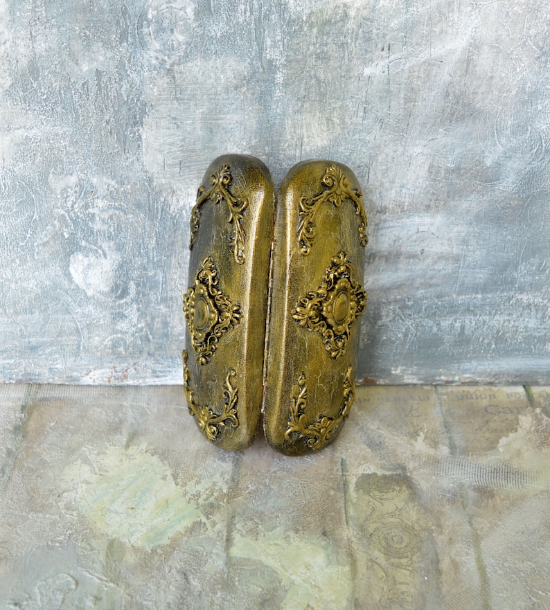 May include: A pair of ornate gold-colored boxes with intricate floral designs and a raised center piece. The boxes are closed and sitting on a textured surface.