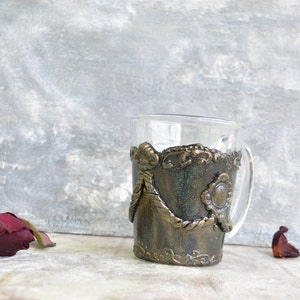 May include: A glass mug with a decorative, textured, gold and green design. The design features a rope-like pattern and a circular, embossed detail.