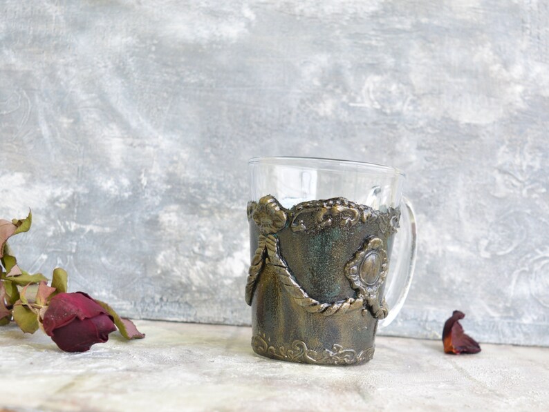 May include: A glass mug with a decorative, textured, gold and green design. The design features a rope-like pattern and a circular, embossed detail.