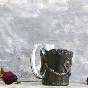 May include: A clear glass mug with a decorative, dark brown, metal-like sleeve. The sleeve has a rope-like design and a large, round, gemstone-like accent.