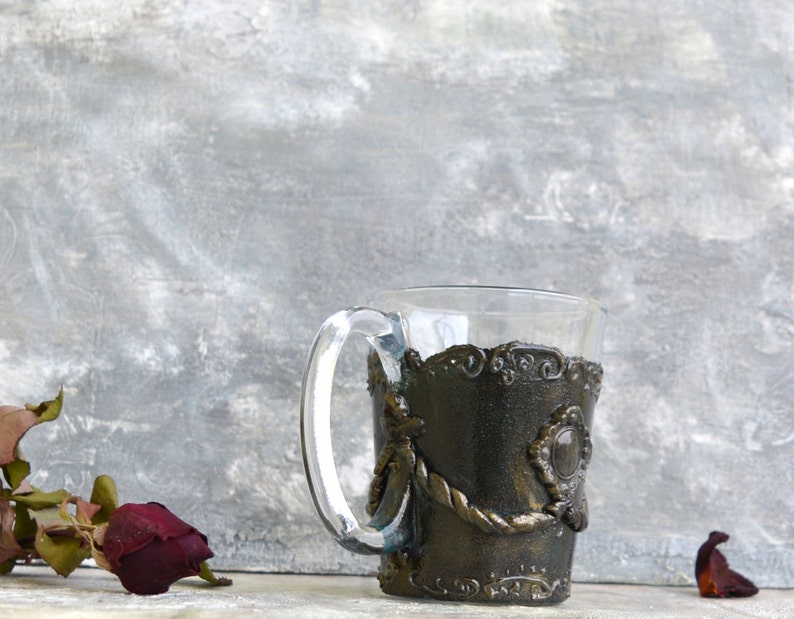 May include: A clear glass mug with a decorative, dark brown, metal-like sleeve. The sleeve has a rope-like design and a large, round, gemstone-like accent.