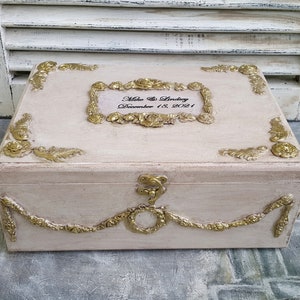 Peut inclure: Une boîte en bois blanche avec des accents dorés et un motif floral décoratif. La boîte a un couvercle à charnières et une serrure. Le couvercle est gravé du texte "Mike & Lindsey December 18, 2021".