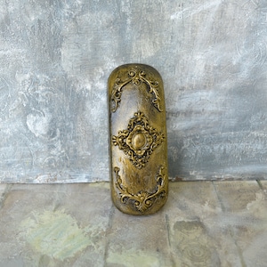 May include: A gold-colored decorative box with an ornate design. The box has a raised, circular design in the center and is surrounded by intricate floral patterns.