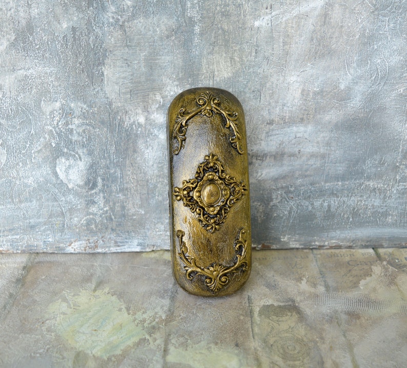 May include: A gold-colored decorative box with an ornate design. The box has a raised, circular design in the center and is surrounded by intricate floral patterns.