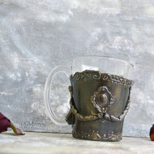 May include: A glass mug with a decorative, metallic, gold and brown, textured, sleeve. The sleeve has a raised, circular design with a smaller, circular, silver-colored accent in the center.