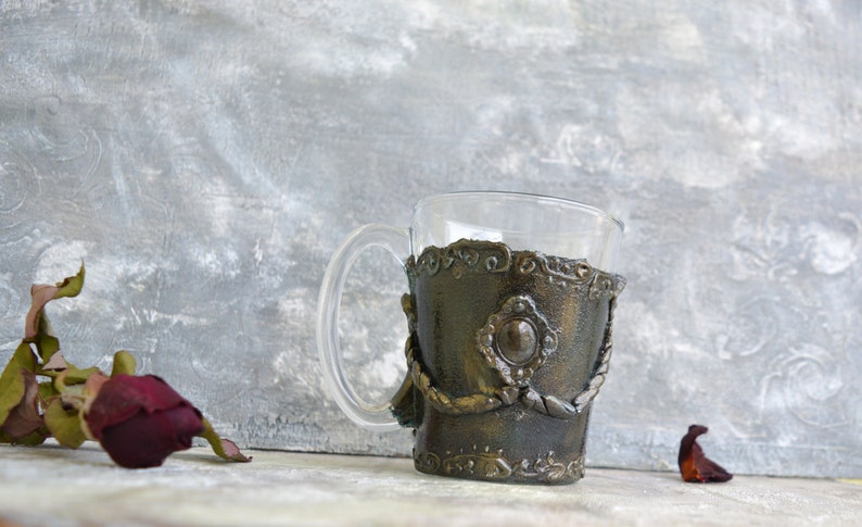 May include: A glass mug with a decorative, metallic, gold and brown, textured, sleeve. The sleeve has a raised, circular design with a smaller, circular, silver-colored accent in the center.