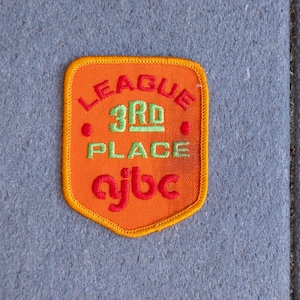 May include: An orange embroidered patch with the words "LEAGUE 3RD PLACE ajbc" in red and green. The patch has a yellow border and is set against a gray background. The patch is likely for a sports team or league.