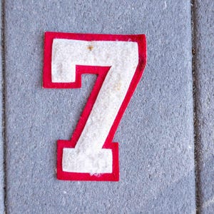 May include: A red and white felt number seven patch. The number is white with a red border. The patch is on a gray surface.