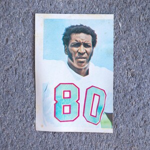 Puede incluir: Tarjeta de fútbol vintage con un jugador con una camiseta blanca con el número 80 en verde azulado con un contorno rosa. El jugador tiene una expresión seria sobre un fondo azul claro.