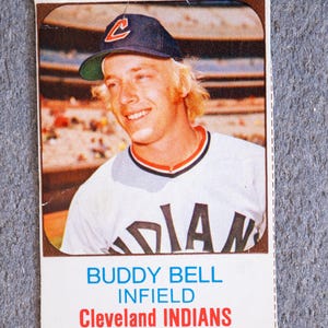 May include: Vintage baseball card featuring Buddy Bell of the Cleveland Indians. The card shows a smiling player wearing a navy blue cap with a red "C" logo and a white jersey with "INDIANS" across the chest. The card has a white border with text.