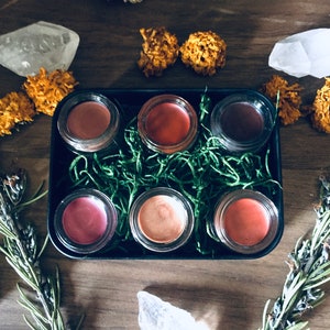 May include: Six small glass jars with lids, filled with different shades of lipstick, arranged in a black tray lined with green moss. The tray is on a wooden surface with crystals and dried flowers.