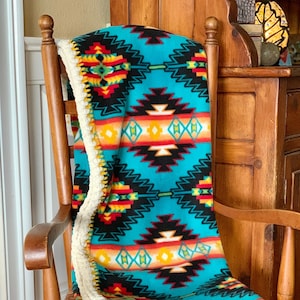 Op de afbeelding: Een turquoise, zwart, rood, geel en groene gepatroneerde fleece deken met een witte gehaakte rand. De deken is over een houten stoel gelegd.