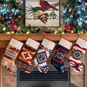 以下が含まれることがあります： 幾何学模様のカラフルな靴下4つが暖炉の棚からぶら下がっています。靴下は赤、青、茶色、白の幾何学模様で飾られています。暖炉は電飾と緑色の花輪で飾られています。