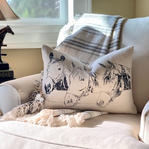 Op de afbeelding: Een witte fauteuil met een crèmekleurige deken en een kussen met een zwart-witte print van drie paardenkoppen.