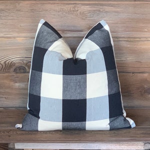 May include: A square pillow with a black, white, and light gray buffalo check pattern. The pillow is resting on a wooden surface. The pillow has a soft, inviting appearance, suitable for home decor.