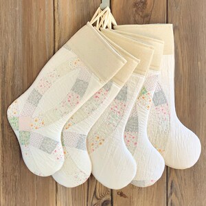 May include: Five white quilted Christmas stockings with a patchwork design featuring pink, green, and gray floral patterns. The stockings are hanging from a hook on a wooden wall.