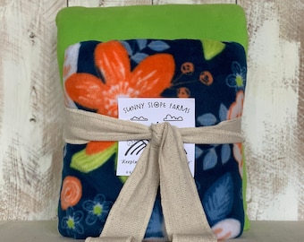 May include: A folded blanket with a floral pattern in orange, blue, and green, tied with a beige ribbon. The blanket is wrapped around a green blanket. The label reads "Sunny Slope Farms".