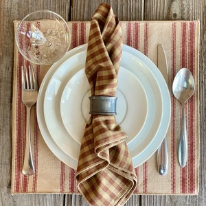 May include: A brown and tan checkered napkin with a silver napkin ring is placed on a white plate with a smaller white plate underneath. The plates are on a beige placemat with red stripes. A silver fork, knife, and spoon are on the placemat.