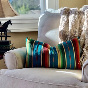 May include: A white armchair with a throw blanket and a colorful striped pillow. The armchair is in a living room setting with a window and a lamp in the background.