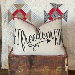 May include: A white pillow with red trim and the text "Let freedom ring" in black cursive lettering with an arrow pointing to the right. The pillow is sitting on top of a wooden box with a floral pattern.