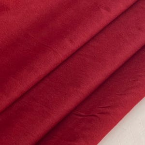 May include: Close-up of folded red fabric. The fabric is a deep, rich shade of red, with subtle variations in texture and tone. The folds create diagonal lines across the image, adding depth and visual interest. The fabric appears soft and possibly luxurious.