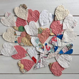 May include: A collection of quilted heart shapes in various colors and patterns. The hearts are made from different fabrics, including white, pink, and patterned textiles. The arrangement is on a white wooden surface.