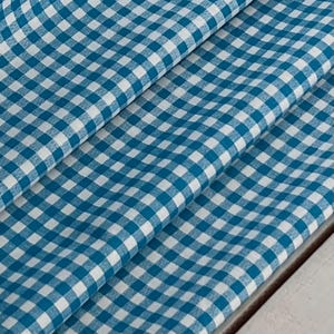 May include: Close-up of a folded fabric with a blue and white checkered pattern. The fabric is draped, showing the texture and the interplay of light and shadow across the surface. The pattern is a classic gingham design.
