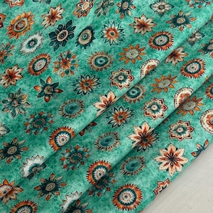 Puede incluir: Primer plano de una tela con un estampado floral repetido. Las flores son en tonos naranja, verde azulado y blanco, sobre un fondo verde moteado. La tela está doblada, mostrando la textura y el diseño.