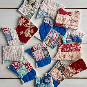 May include: A collection of small, irregularly shaped fabric pieces, likely quilt blocks, in various colors and patterns. The pieces feature floral, geometric, and patchwork designs, with a mix of red, blue, white, and patterned fabrics. They are arranged on a white wooden surface.