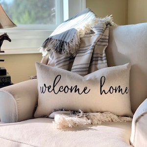 May include: A white throw pillow with black text that reads "welcome home" on a beige armchair with a striped throw blanket draped over the back.