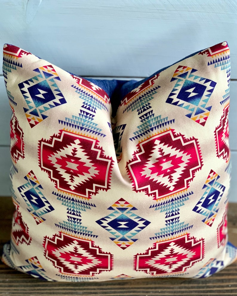 May include: A decorative pillow with a geometric pattern in shades of red, blue, and yellow on a cream background. The pillow has a denim-like fabric backing.