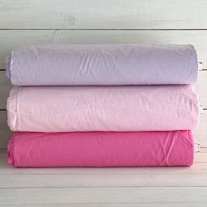 May include: Three rolls of fabric in shades of purple, pink, and fuchsia are stacked on top of each other. The fabric appears to be a soft material, and the rolls are neatly folded. The background is a white wooden surface.