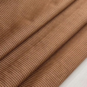 Puede incluir: Tela rayada marrón y beige con un sutil patrón de ondas. La tela está doblada por la mitad, mostrando la textura y el tejido.