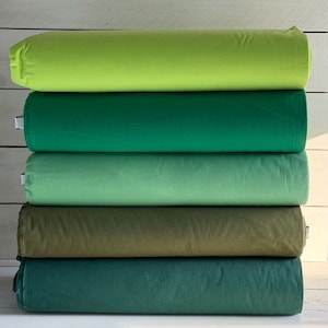 May include: A stack of five fabric rolls in varying shades of green. The colors range from a bright lime green at the top, transitioning through emerald, mint, olive, and ending with a deep teal at the bottom. The rolls are neatly stacked.