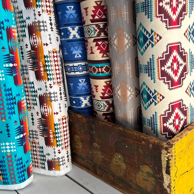 May include: A collection of six fabric bolts with colorful geometric patterns. The fabrics are in various shades of blue, red, orange, and white. The bolts are stacked on top of each other and are sitting on a wooden crate.