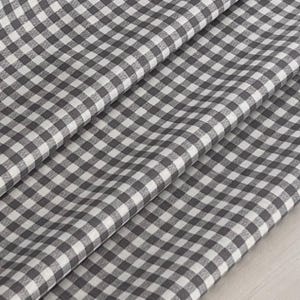 May include: Close-up of a gray and white gingham fabric. The fabric is folded, showing multiple layers of the checkered pattern. The pattern consists of evenly spaced squares in a classic gingham design, creating a textured appearance.