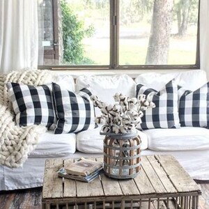 May include: A cozy living room scene featuring a white sofa adorned with black and white buffalo check pillows and a chunky knit blanket. A rustic wooden coffee table holds books and a vase of cotton stems. A floor lamp and window complete the farmhouse aesthetic.