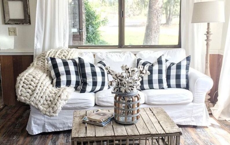 May include: A cozy living room scene featuring a white sofa adorned with black and white buffalo check pillows and a chunky knit blanket. A rustic wooden coffee table holds books and a vase of cotton stems. A floor lamp and window complete the farmhouse aesthetic.