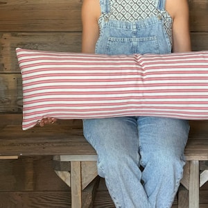 May include: A long, rectangular pillow with a red and white striped pattern. The pillow is held by a person wearing denim overalls. The pillow's design is reminiscent of classic farmhouse decor.