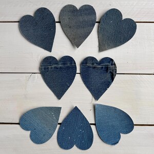 May include: Nine denim heart shapes in varying shades of blue are arranged on a white wooden surface. The hearts appear to be cut from recycled denim fabric, showcasing the texture and color variations of the material. Ideal for crafting or decor.