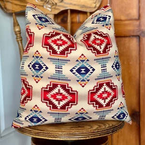 May include: A decorative throw pillow with a Southwestern-inspired geometric pattern in shades of red, blue, and beige. The pillow is sitting on a wooden chair.