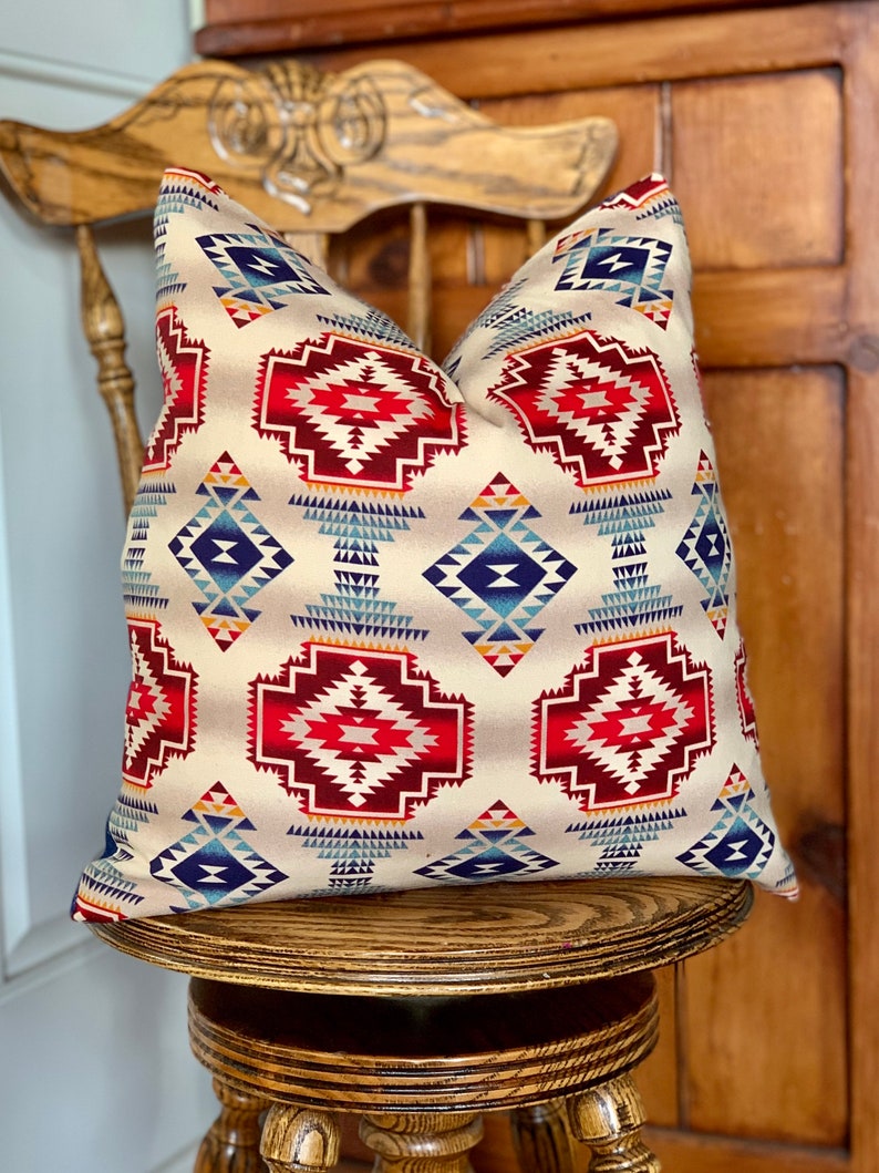 May include: A decorative throw pillow with a Southwestern-inspired geometric pattern in shades of red, blue, and beige. The pillow is sitting on a wooden chair.