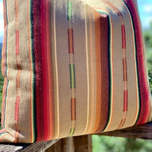 May include: A decorative pillow with a woven fabric cover. The pillow features vertical stripes in shades of red, brown, green, and tan. The pillow is resting on a wooden surface, with a blurred background of greenery and a blue sky.