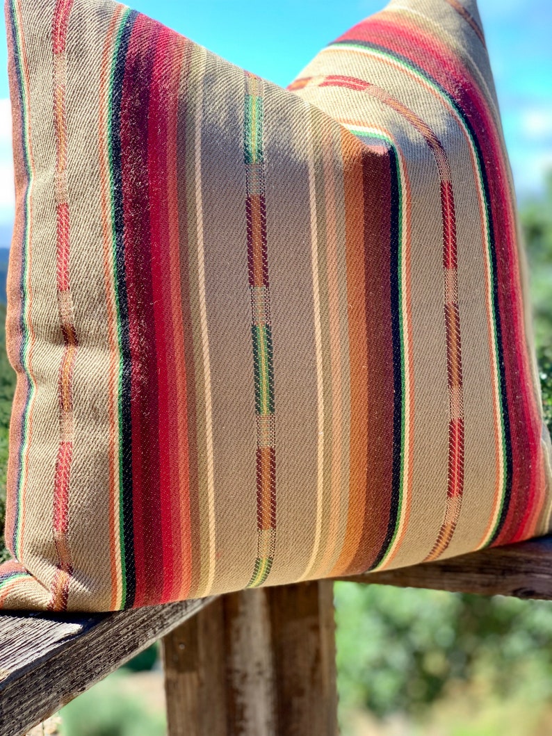 May include: A decorative pillow with a woven fabric cover. The pillow features vertical stripes in shades of red, brown, green, and tan. The pillow is resting on a wooden surface, with a blurred background of greenery and a blue sky.