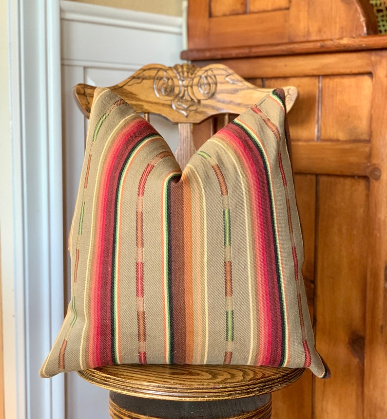 May include: A decorative pillow with a striped pattern in shades of tan, red, brown, and green. The pillow is placed on a wooden chair with a carved back. The pillow's design features vertical stripes.