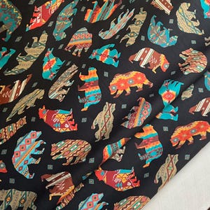 Op de afbeelding: Zwarte stof met een herhaald patroon van kleurrijke berensilhouetten. De beren hebben geometrische ontwerpen in rood, oranje, turquoise en bruin. De stof is waarschijnlijk katoen of een vergelijkbaar materiaal.