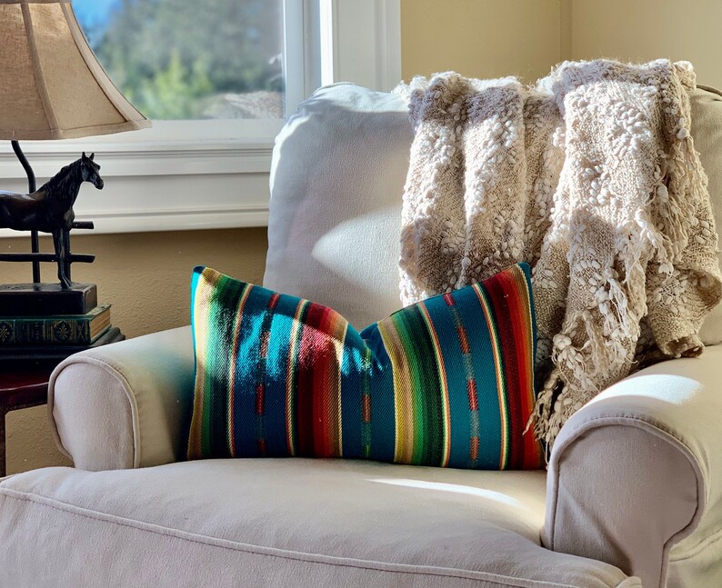 May include: A white armchair with a throw blanket and a colorful striped pillow. The armchair is in a living room setting with a window and a lamp in the background.